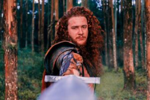 A man in the forest holding a sword menacingly with the point held near the viewers eyes.